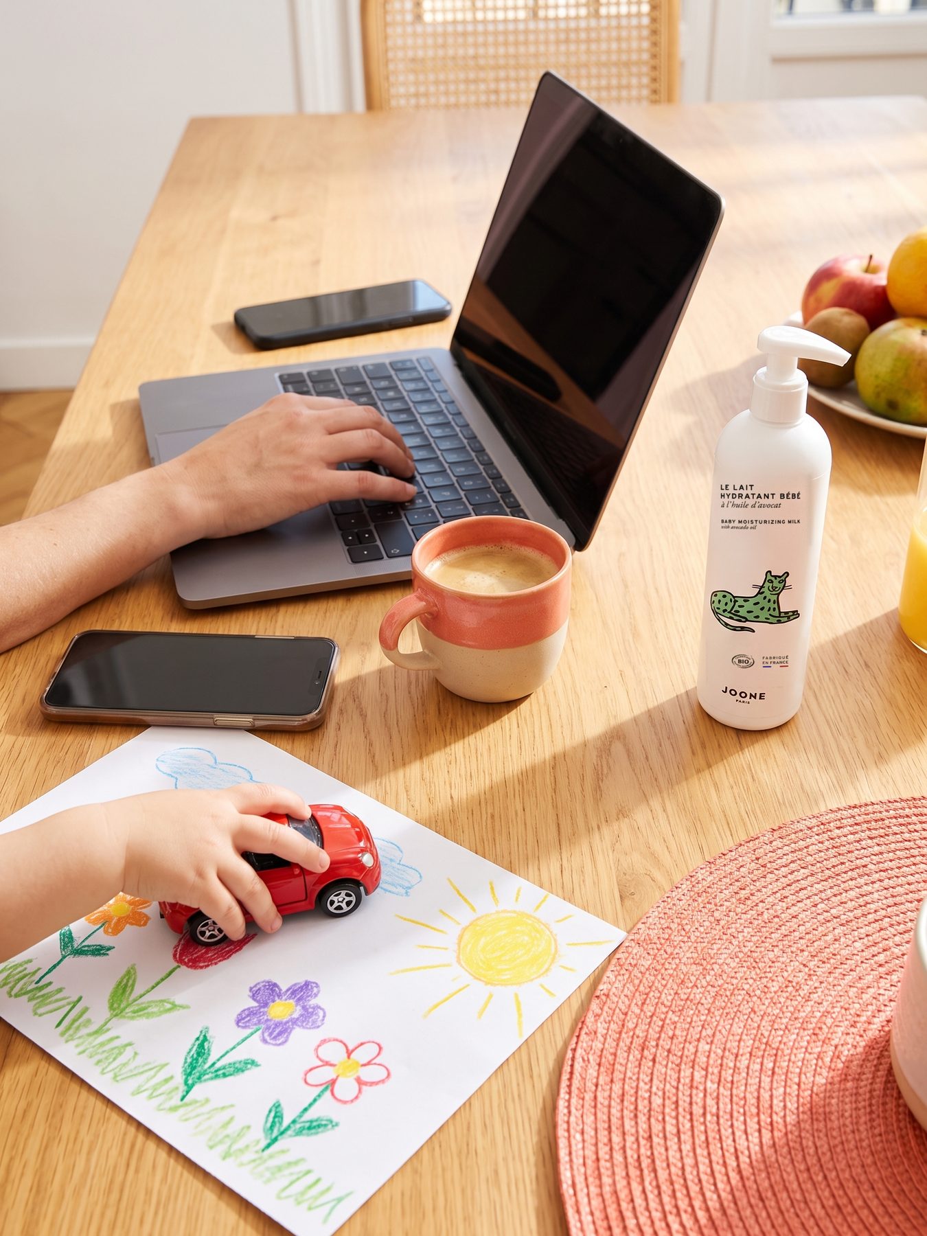 keukentafel met laptop, koffie, joone-fles, een kindertekening en een kinderhand met een speelgoedauto